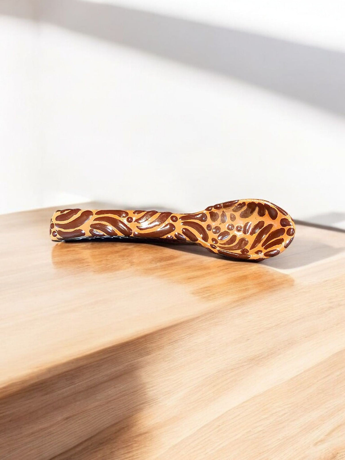 The Vela Lady handcrafted brown Talavera spoon with intricate painted patterns resting on a wooden surface.