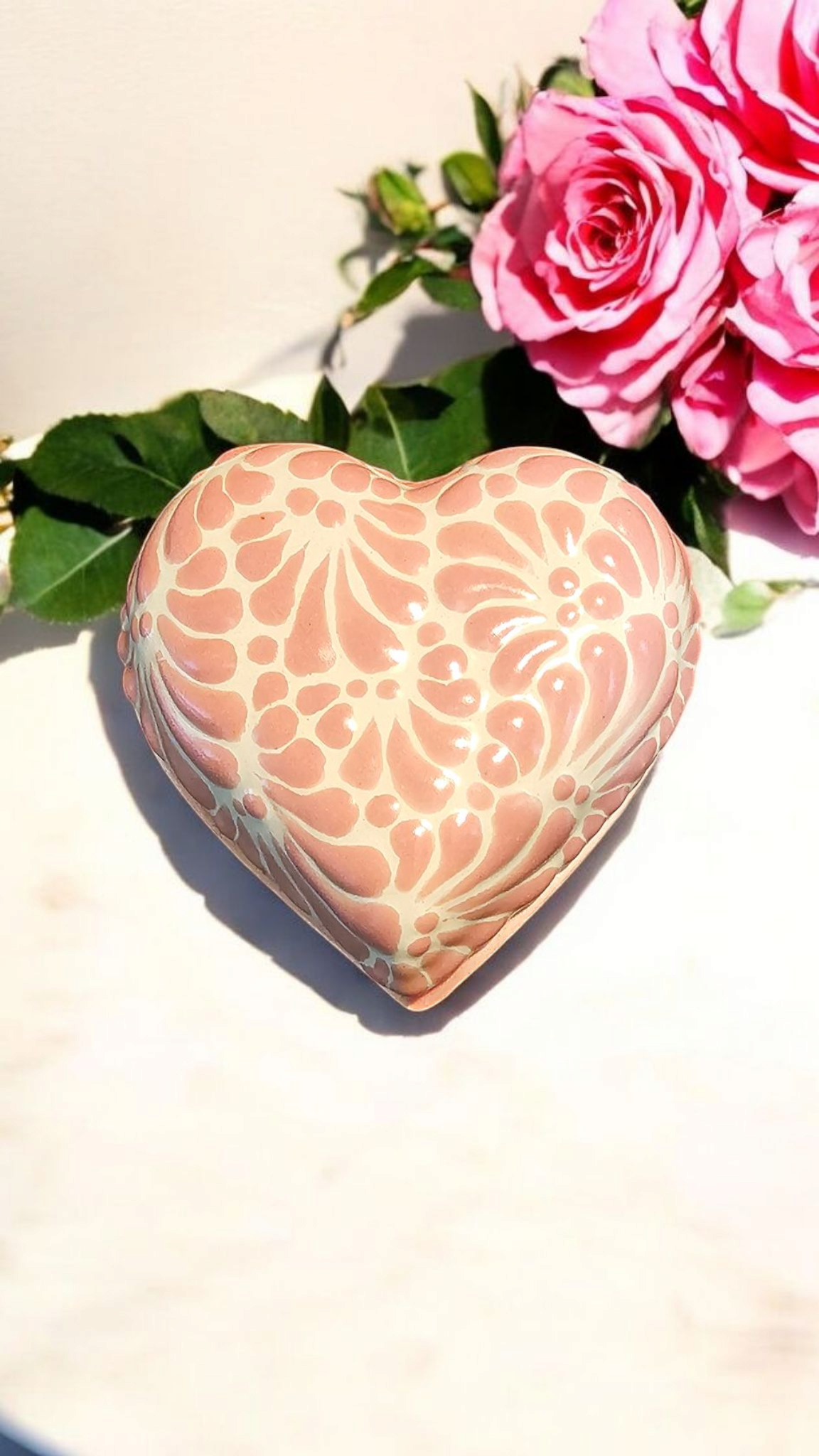 The Vela Lady heart-shaped pink and white Talavera ceramic trinket holder with floral patterns next to pink roses.
