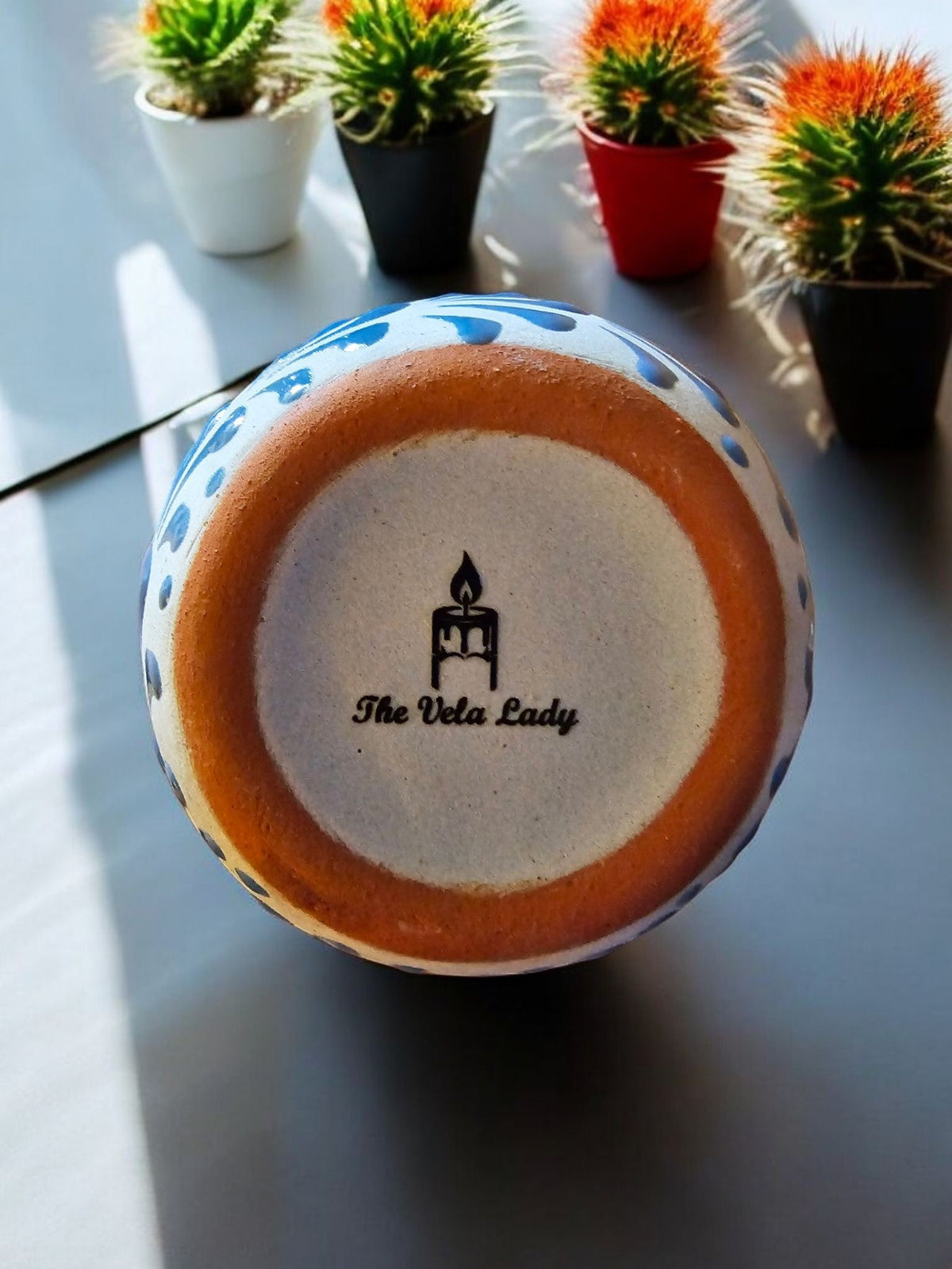 The Vela Lady blue hand-painted Mexican barro mug candle with brand logo on the bottom, set near small cacti.