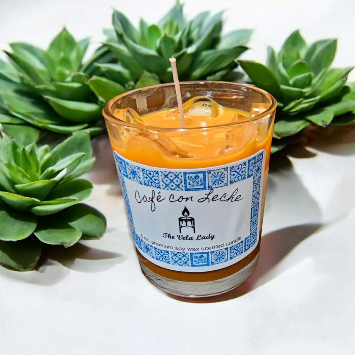 The Vela Lady Cafe con Leche soy wax candle in a glass jar, surrounded by green succulent plants.