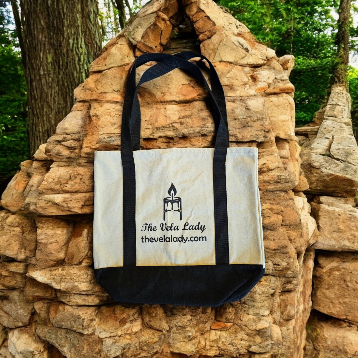 The Vela Lady reusable canvas tote bag in natural and black with a candle logo, displayed against a stone background.