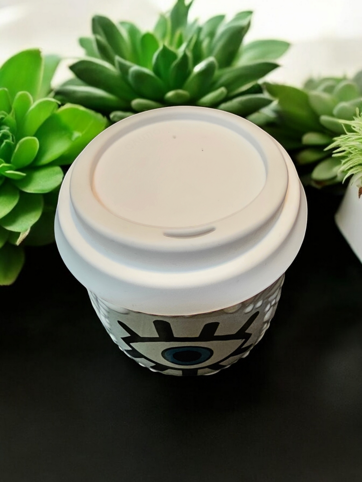 The Vela Lady white Talavera tumbler candle with an evil eye design, topped with a white lid, set against greenery.