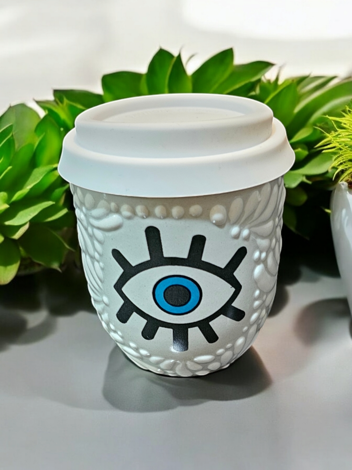 The Vela Lady white Talavera tumbler candle featuring a blue and black evil eye design with a white silicone lid.
