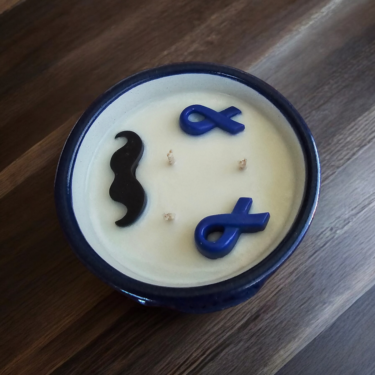 The Vela Lady Men's Health Awareness soy candle in a blue Talavera pottery vessel with mustache and ribbon wax toppers.