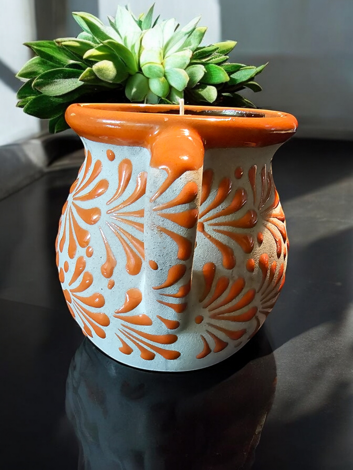 The Vela Lady orange and cream hand-painted Mexican barro mug candle with a succulent plant on a reflective surface.