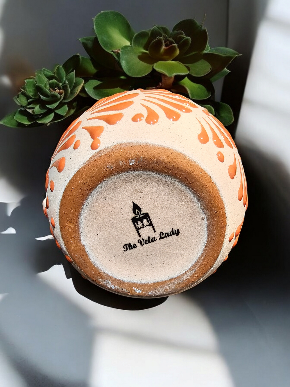 The Vela Lady orange hand-painted Mexican barro mug candle with a succulent plant in the background.