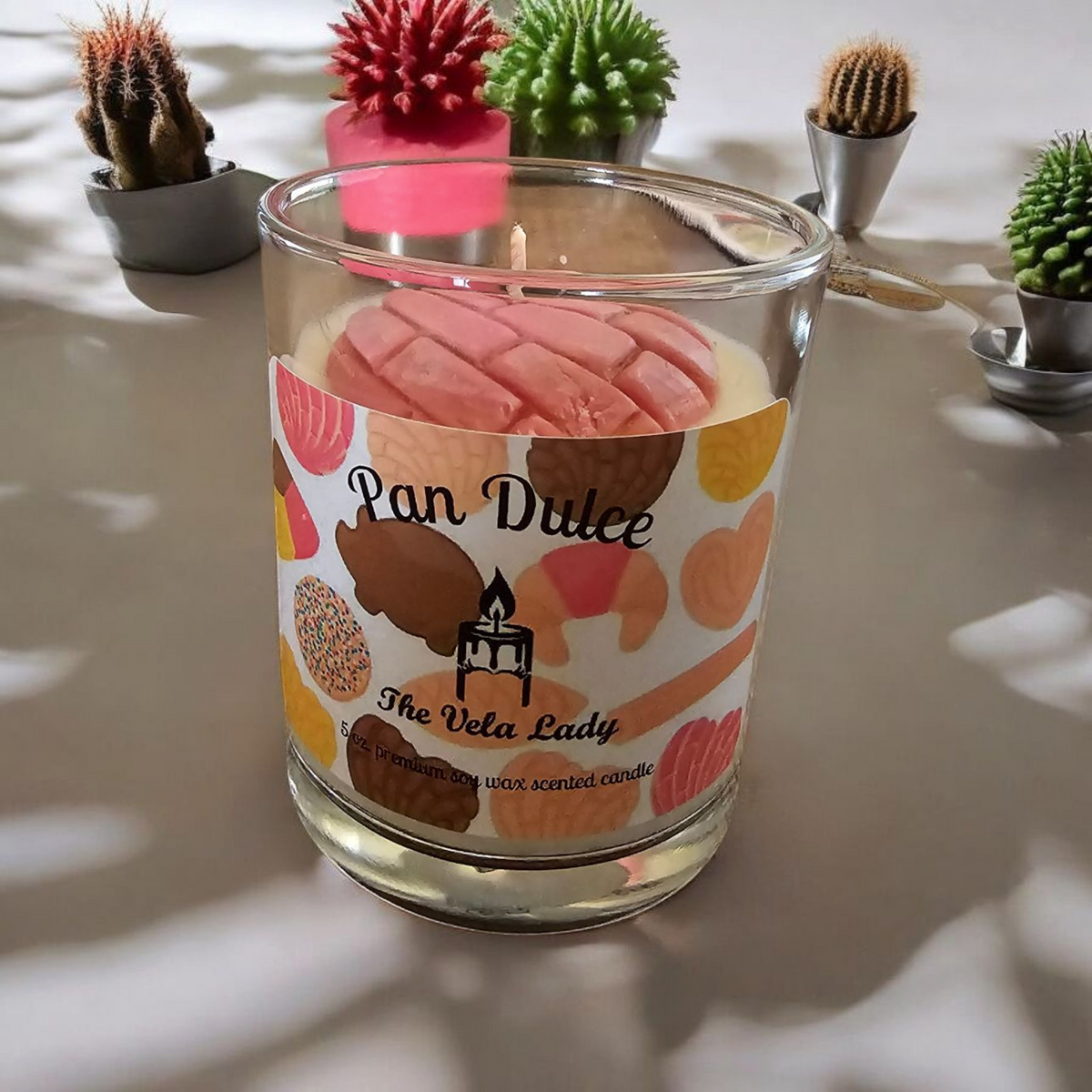 The Vela Lady Pan Dulce soy wax candle in a glass jar with a pink concha-shaped wax topper, surrounded by mini cacti.