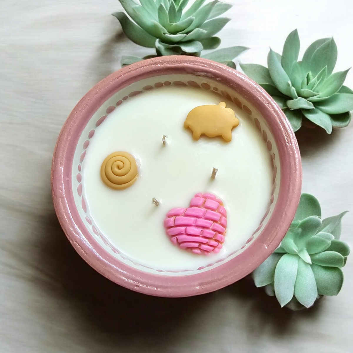 The Vela Lady pink Talavera salsa bowl candle with pan dulce wax embeds and three wicks, surrounded by succulents.