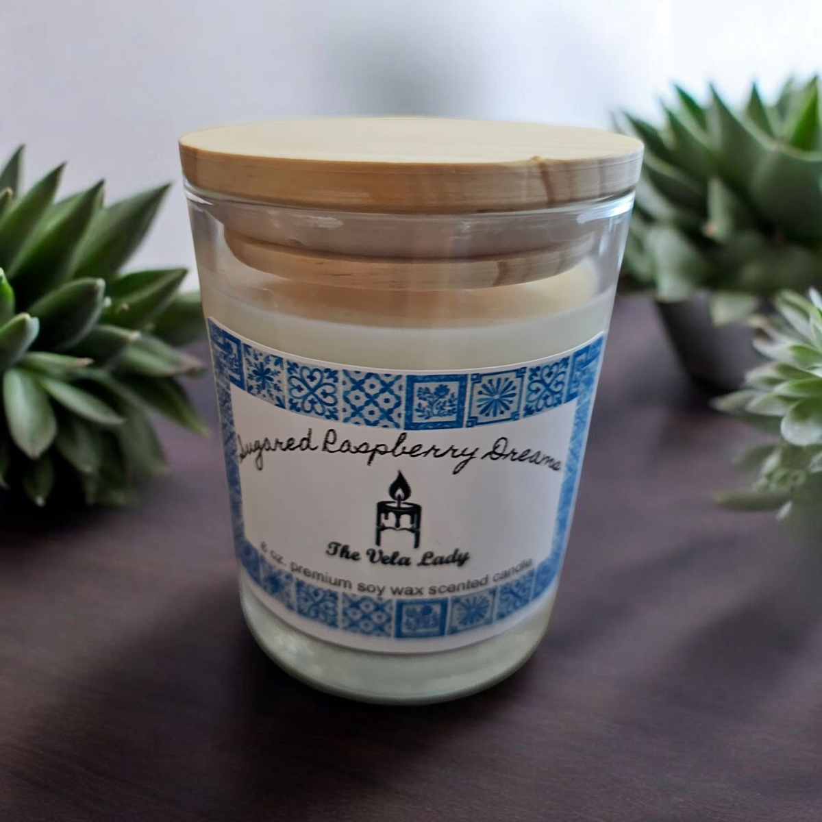 The Vela Lady soy wax candle in a glass jar with a wooden lid and a blue patterned label on a dark surface.