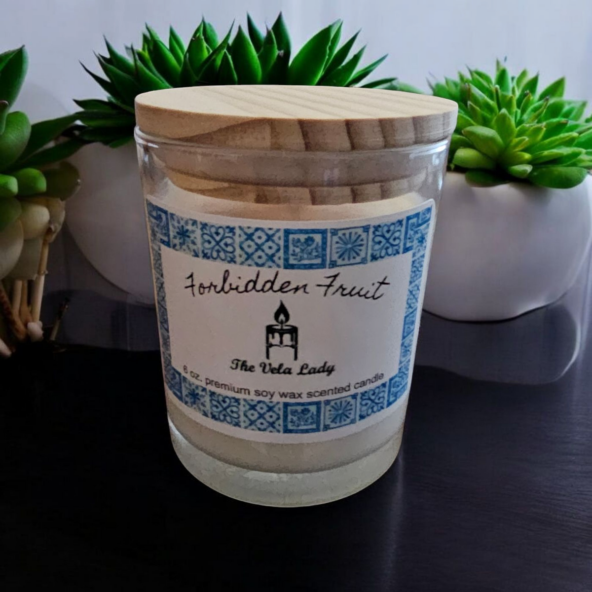 The Vela Lady soy wax candle in a glass jar with a wooden lid and Forbidden Fruit label, set against green plants.