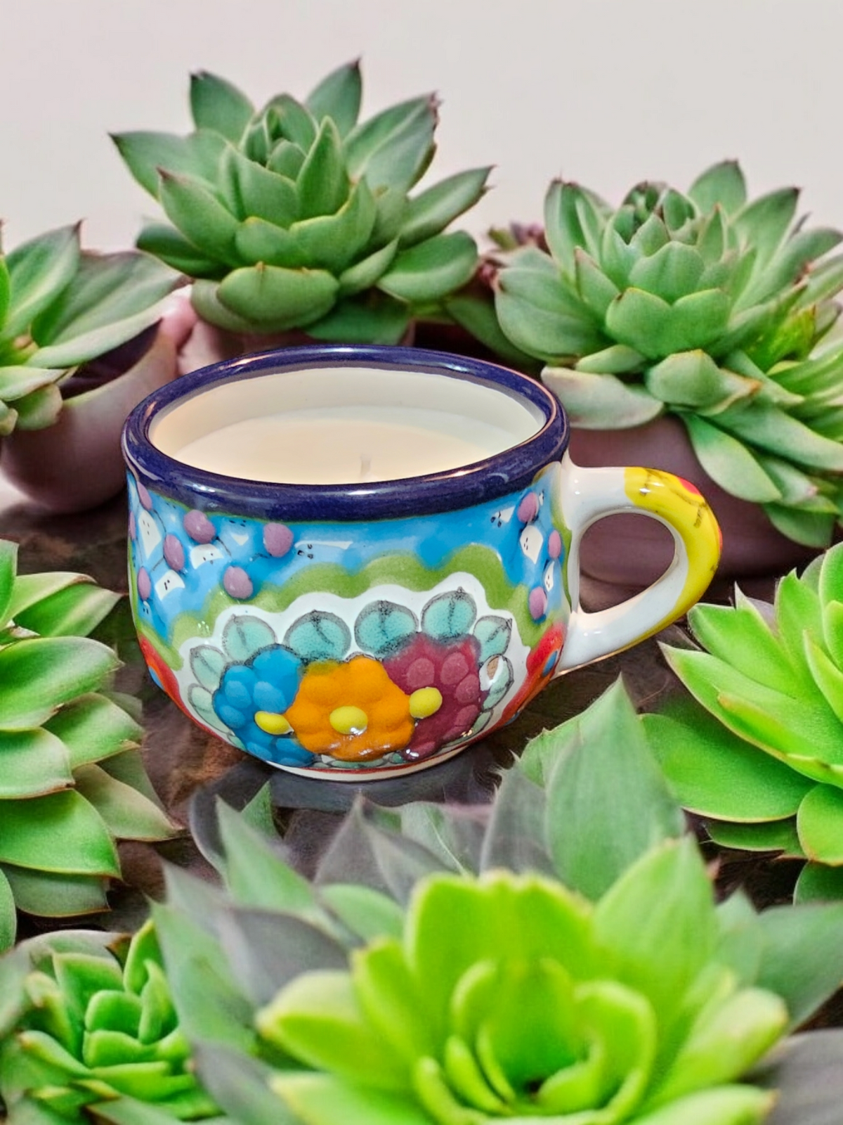 The Vela Lady La Catrina Talavera mug candle with vibrant floral patterns, surrounded by green succulent plants.