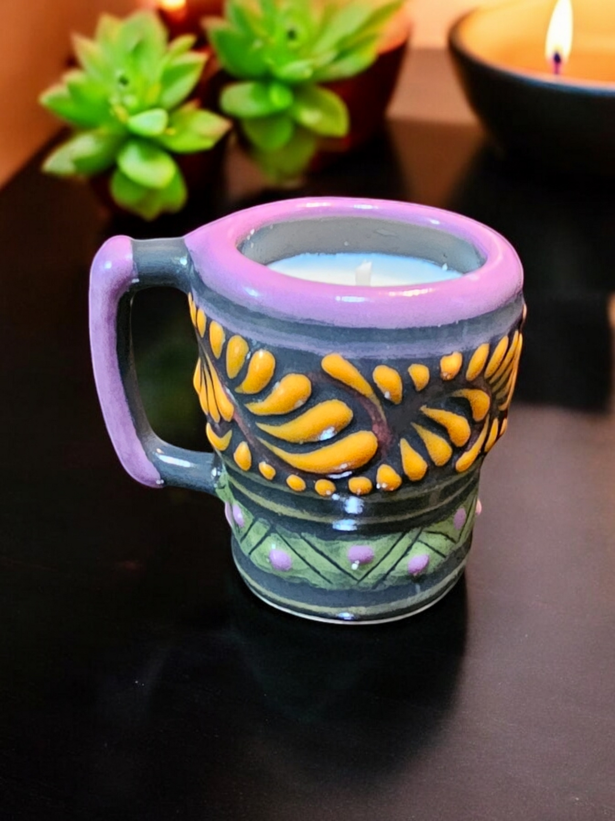 The Vela Lady Calavera shot glass candle with purple rim and yellow floral Talavera patterns on a black surface.