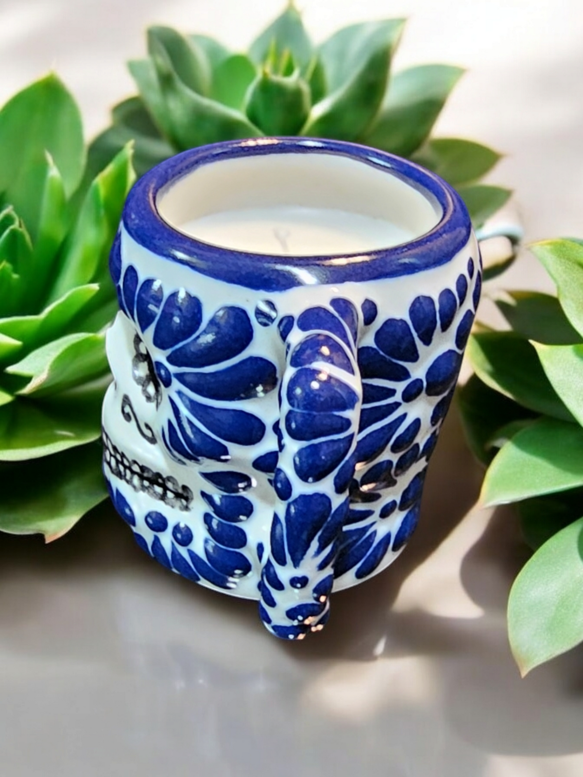 The Vela Lady Calavera shot glass candle with blue and white floral Talavera patterns, set against green succulents.