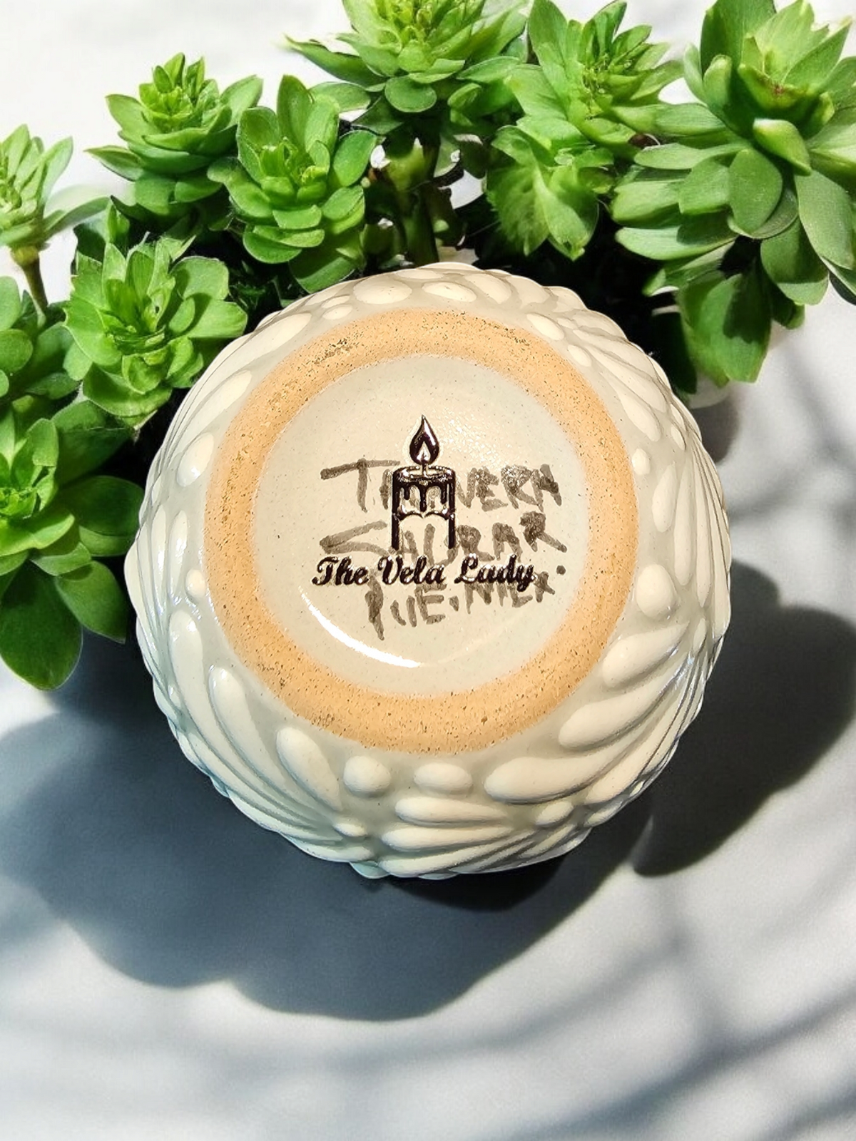 The Vela Lady Buenas Vibras white Talavera ceramic candle vessel bottom with artisan branding and green foliage